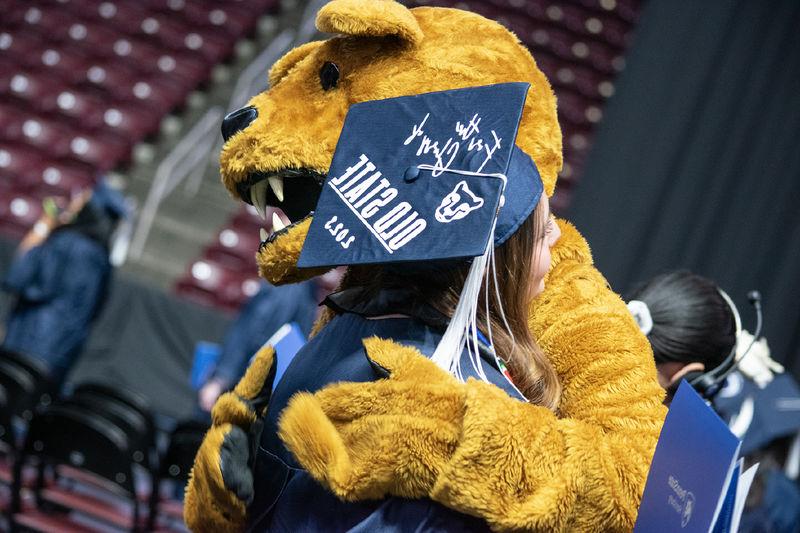 The Nittany Lion hugs a graduate in a cap that reads "For the Glory of Old State 2023"